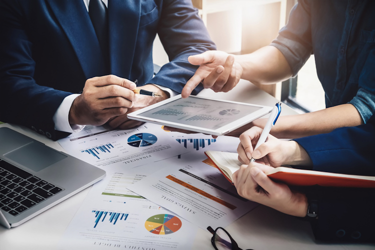 Businessman pointing to tablet with pie chart and bar graphs with two individuals taking notes and analyzing numbers