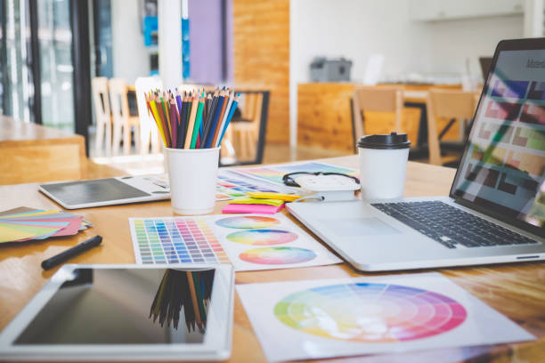 Desktop with white cup full of colored pencils with a modern laptop and tablet, plus printed color wheels laid out 